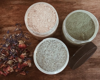 May include: Three glass jars filled with different colored powders. The jars are on a wooden surface. The powders are light pink, green, and light gray.