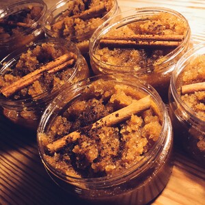 May include: Close-up of six glass jars filled with brown sugar scrub and cinnamon sticks. The jars are arranged in a row on a wooden surface.