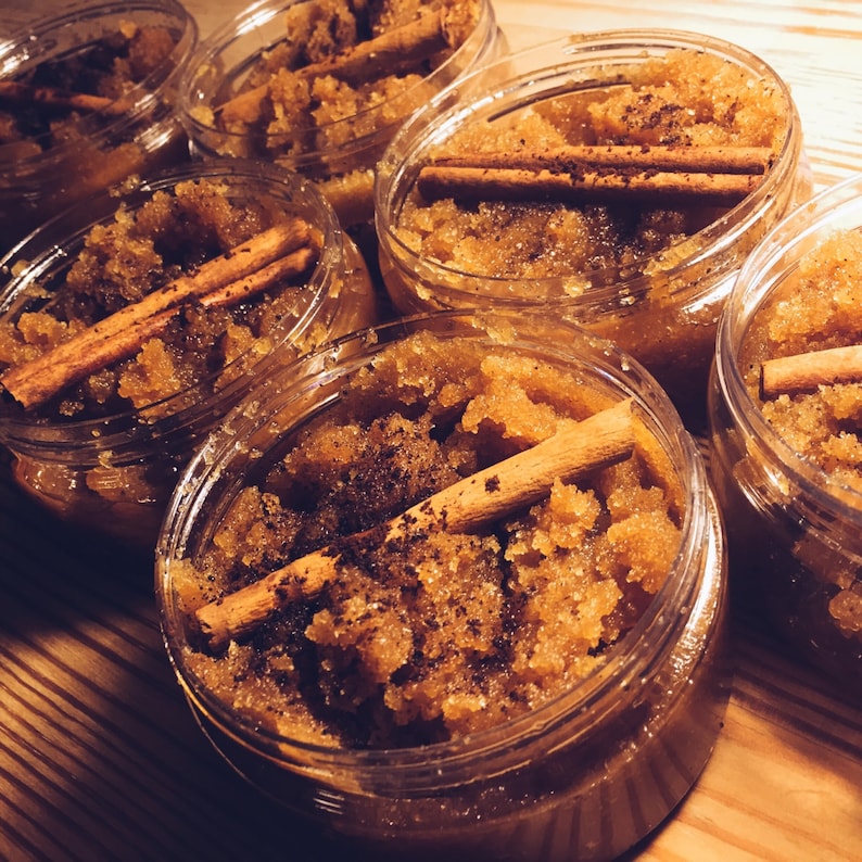 May include: Close-up of six glass jars filled with brown sugar scrub and cinnamon sticks. The jars are arranged in a row on a wooden surface.