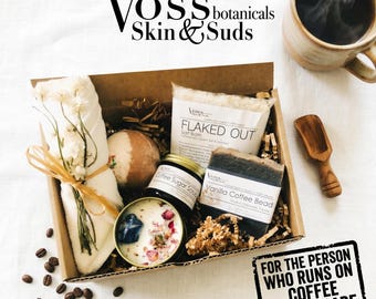 May include: A gift box with Voss Botanicals products, including a candle, soap, bath salts, and a coffee mug. The box contains a variety of self-care items, with the text "FOR THE PERSON WHO RUNS ON COFFEE + SELF-CARE."