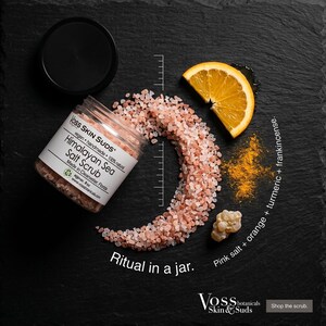 Puede incluir: Un tarro de exfoliante de sal marina del Himalaya con tapa negra, rodeado de sal rosa, una rodaja de naranja y cúrcuma. El tarro está etiquetado como "Voss Skin Suds" y "Ritual in a jar."