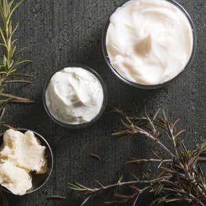 Peut inclure: Vue aérienne de produits de soin. Deux petits bols en verre contiennent de la crème blanche, dont un avec des points verts. Un petit bol en métal contient des morceaux d'une substance solide blanc cassé. Des brins de romarin sont disposés autour des bols sur une surface gris foncé.