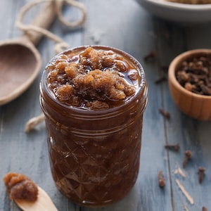 May include: A glass jar filled with brown sugar scrub, with a wooden spoon and a bowl of cloves in the background.