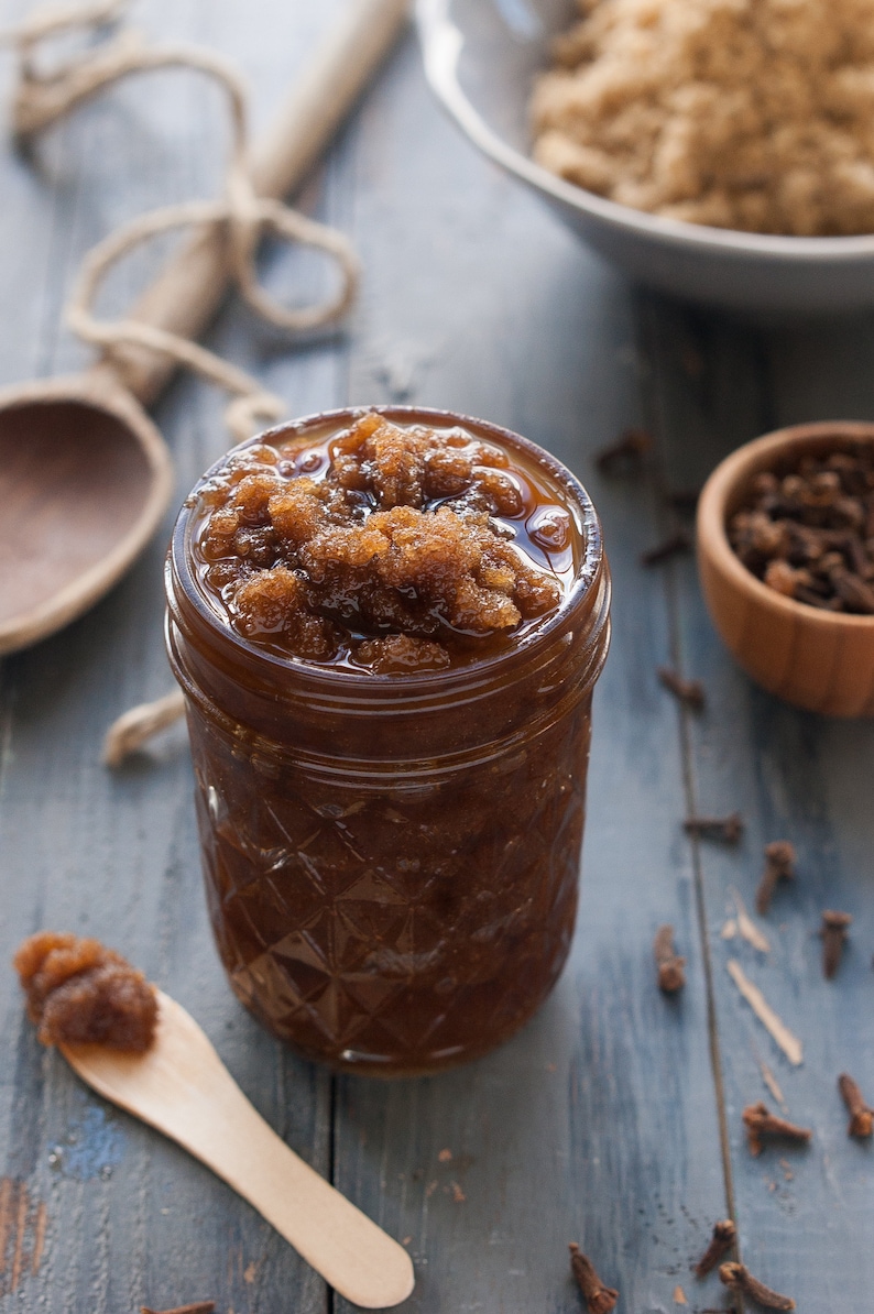 May include: A glass jar filled with brown sugar scrub, with a wooden spoon and a bowl of cloves in the background.