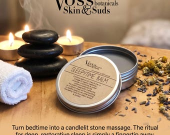 Może przedstawiać: Okrągła metalowa puszka z balsamem Sleepytime Balm marki Voss Botanicals z beżową etykietą i czarnym tekstem. Puszka jest otwarta, ukazując zawartość. Obok znajdują się trzy ułożone w stos czarne kamienie, biały zwinięty ręcznik i suszone kwiaty.