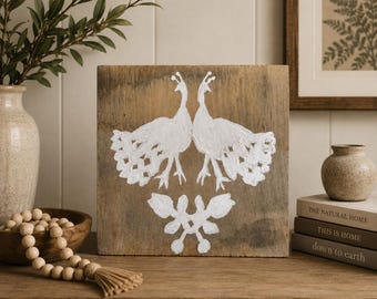 Könnte beinhalten: Ein quadratisches Holzschild mit einem weiß aufgemalten Design von zwei einander zugewandten Pfauen mit gefächerten Schwänzen über einem symmetrischen Blumenmotiv. Das Holz hat eine natürliche, verwitterte braune Oberfläche.