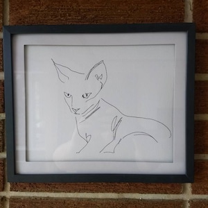K&ouml;nnte beinhalten: Ein gerahmtes Wandbild mit einer schwarzen Tuschezeichnung einer Katze auf wei&szlig;em Hintergrund. Das Kunstwerk befindet sich in einem einfachen schwarzen rechteckigen Rahmen und zeigt einen durchgehenden Linienstil, der Kopf und K&ouml;rper der Katze umrei&szlig;t.