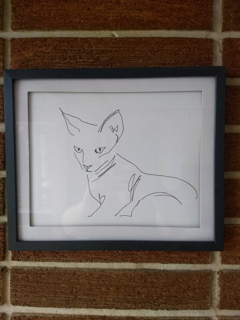 K&ouml;nnte beinhalten: Ein gerahmtes Wandbild mit einer schwarzen Tuschezeichnung einer Katze auf wei&szlig;em Hintergrund. Das Kunstwerk befindet sich in einem einfachen schwarzen rechteckigen Rahmen und zeigt einen durchgehenden Linienstil, der Kopf und K&ouml;rper der Katze umrei&szlig;t.