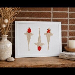 May include: A rectangular white frame containing a print of three koi fish. The fish are depicted in a light beige color with distinct red markings on their heads, arranged vertically against a plain white background within the frame.