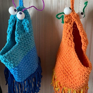 May include: Two handmade, whimsical crochet wall pockets. One is blue with a fringe of dark blue yarn, the other is orange with a yellow fringe. Both have large googly eyes and pipe cleaner antennae, hanging from black hooks.
