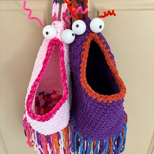 May include: Two whimsical, handmade crochet storage baskets. One is pink and white with pink, orange, and purple fringe, and the other is purple with orange trim and blue fringe. Both have large googly eyes and pipe cleaner antennae.
