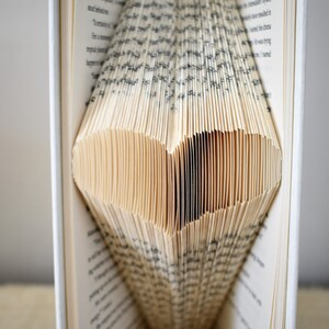 May include: A book with pages folded into a heart shape. The pages are cream colored and the text is black. The book is open and the heart shape is visible in the center of the book.