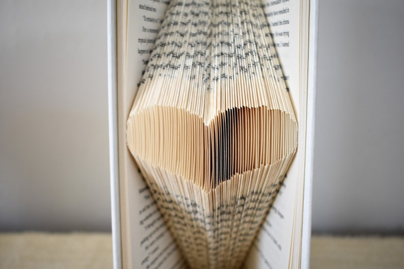 May include: A book with pages folded into a heart shape. The pages are cream colored and the text is black. The book is open and the heart shape is visible in the center of the book.