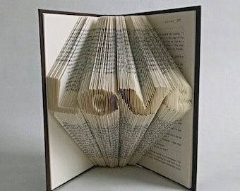 May include: An open book with pages folded to spell the word "LOVE". The book has a dark brown cover and the pages are a light cream color. The word "LOVE" is prominently displayed in the center of the book.