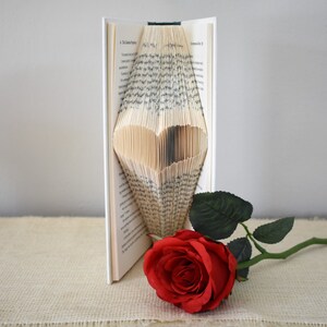 May include: A book with pages folded into a heart shape, standing on a beige surface. A single red rose with green leaves lies next to the book.