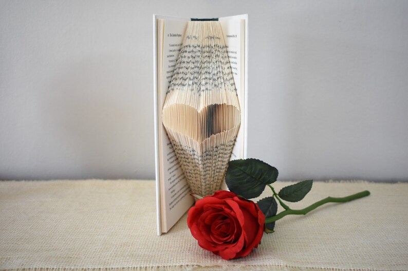 May include: A book with pages folded into a heart shape, standing on a beige surface. A single red rose with green leaves lies next to the book.