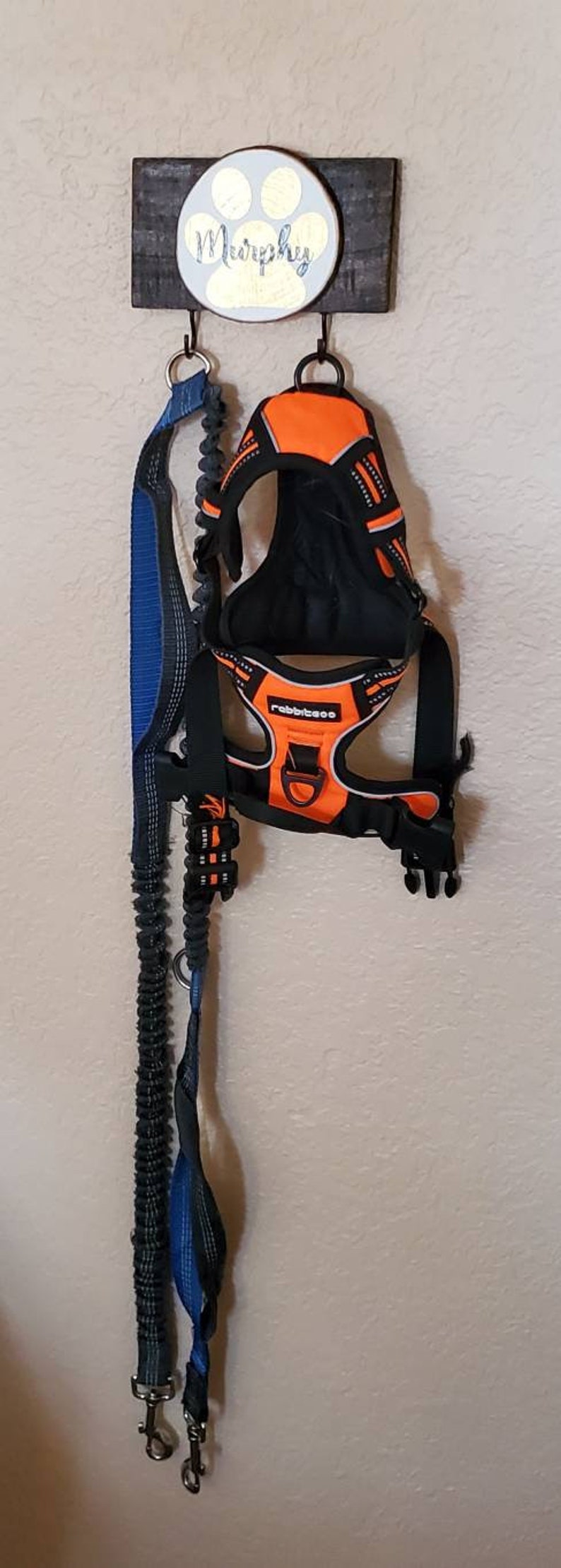 May include: A wooden dog leash and harness holder with a paw print and the name "Murphy" painted on it. A blue leash and an orange harness are hanging from the hooks.