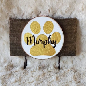May include: A wooden dog leash holder with a gold paw print and the name "Murphy" written in black. The holder has two hooks for hanging leashes.