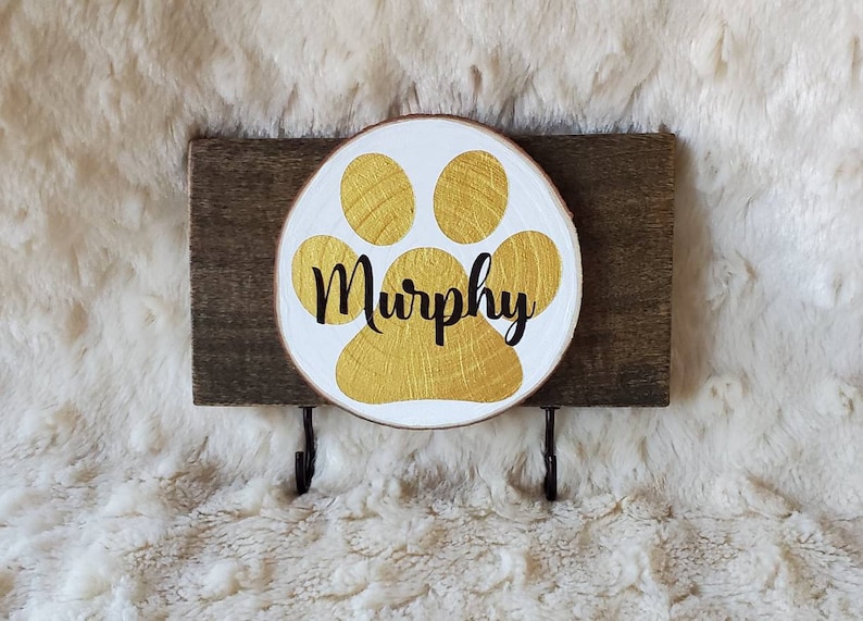 May include: A wooden dog leash holder with a gold paw print and the name "Murphy" written in black. The holder has two hooks for hanging leashes.