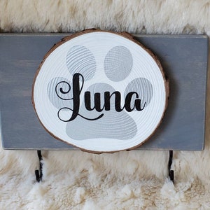 May include: A gray wooden sign with a white wooden paw print and the name "Luna" written in black cursive. The sign has two black metal hooks on the bottom.