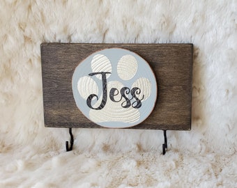 May include: A wooden sign with a light blue paw print and the name "Jess" written in black. The sign has two black metal hooks on the bottom.