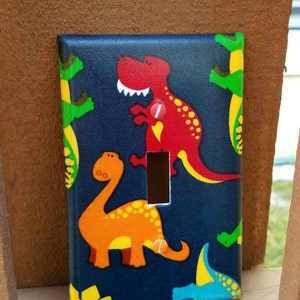 May include: A blue light switch cover with a dinosaur pattern. The cover features a red dinosaur, an orange dinosaur, and a blue dinosaur. The dinosaurs are on a dark blue background with yellow and green accents.