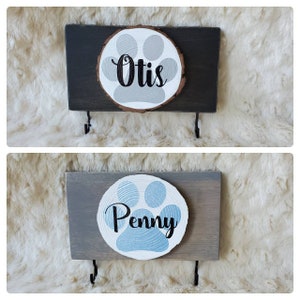 May include: Two wooden dog leash holders with paw print designs. The top holder is black with a gray paw print and the name "Otis" written in black. The bottom holder is gray with a blue paw print and the name "Penny" written in black.