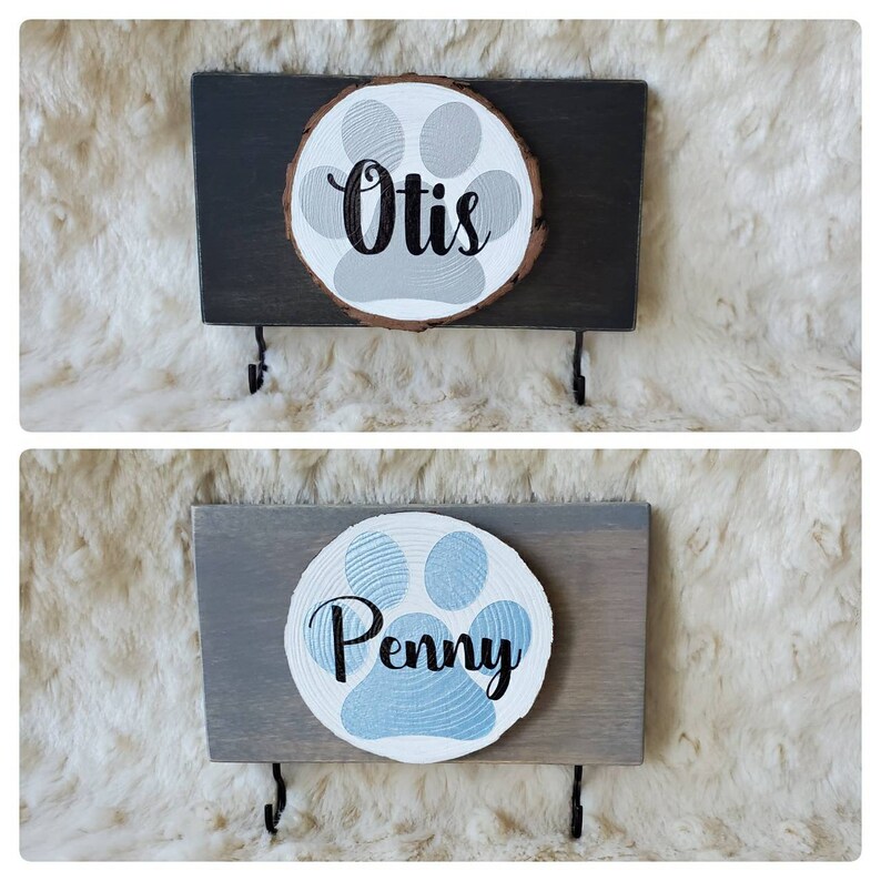 May include: Two wooden dog leash holders with paw print designs. The top holder is black with a gray paw print and the name "Otis" written in black. The bottom holder is gray with a blue paw print and the name "Penny" written in black.