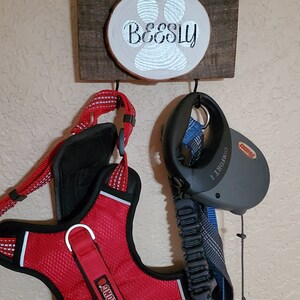 May include: A wooden sign with a paw print and the name "Beesly" on it. Two hooks are attached to the sign, one holding a red dog harness with a Kong logo and the other holding a black retractable dog leash.