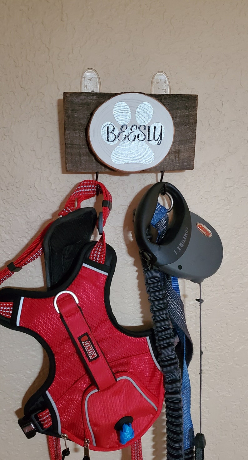 May include: A wooden sign with a paw print and the name "Beesly" on it. Two hooks are attached to the sign, one holding a red dog harness with a Kong logo and the other holding a black retractable dog leash.