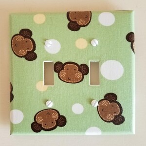 May include: A light green light switch cover with a white polka dot pattern and brown monkey faces. The cover has two light switch openings.