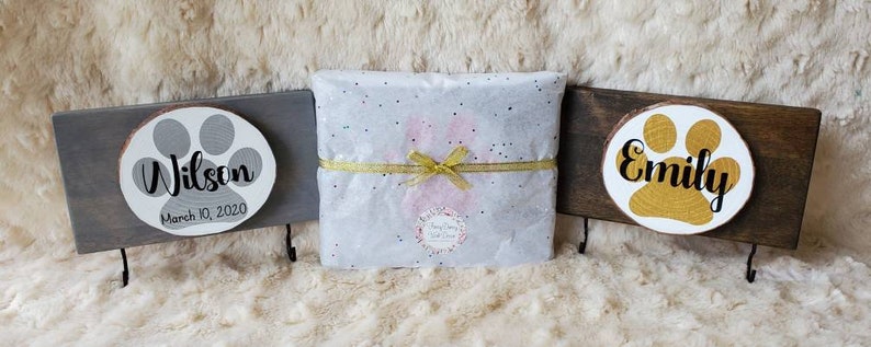 May include: Three wooden pet memorial signs with paw prints and names. The signs are on a white fuzzy surface. The signs are for Wilson, Emily, and a third pet. The signs have hooks on the bottom.