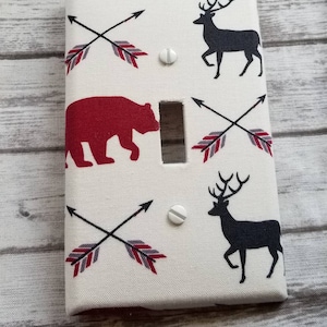 May include: A white light switch cover with a repeating pattern of black deer, red bears, and crossed arrows with red and gray feather details. The cover has two white screws.