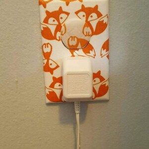 Puede incluir: Una tapa de tomacorriente blanca con un dise&ntilde;o de impresi&oacute;n de zorro. La tapa tiene un adaptador el&eacute;ctrico blanco enchufado.