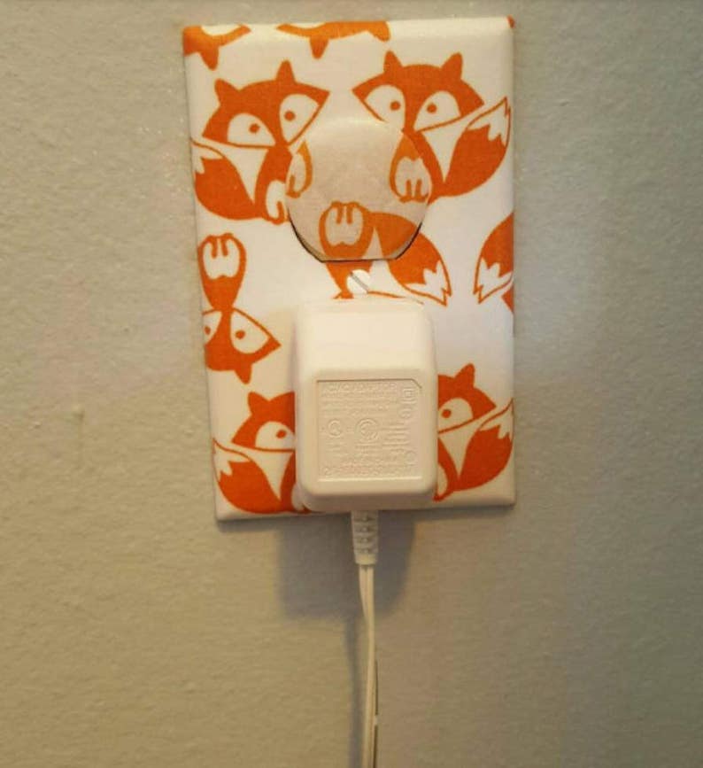 Puede incluir: Una tapa de tomacorriente blanca con un dise&ntilde;o de impresi&oacute;n de zorro. La tapa tiene un adaptador el&eacute;ctrico blanco enchufado.