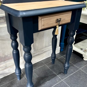 May include: A dark blue wooden side table with a single drawer and turned legs. The drawer has a light wood finish and a decorative brass knob with a tassel. The table is sitting on a dark grey tiled floor.