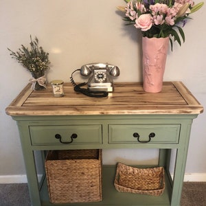 May include: A green console table with two drawers and a lower shelf. The table has a wood top and is decorated with a vintage rotary phone, a candle, and a vase of flowers.