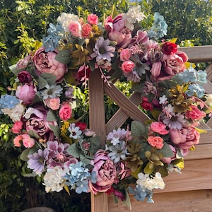 May include: A vibrant floral wreath featuring an array of colorful blooms. The wreath is adorned with large peonies, roses, and smaller flowers in shades of pink, purple, blue, and white, with green foliage. The wreath is displayed on a wooden fence.