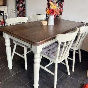 May include: A white farmhouse style dining table with a dark brown wood top and four white chairs. The chairs have a slatted back and are upholstered in grey fabric. A vase of flowers sits on the table.