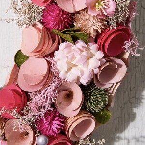 May include: A floral wreath featuring a variety of pink and red flowers, including layered wooden blooms, spherical accents, and delicate sprigs. The arrangement is set against a weathered white wooden background, creating a rustic and charming aesthetic.