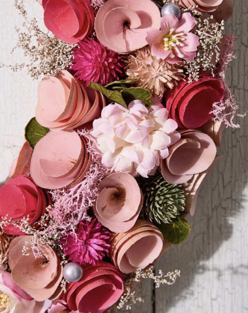 May include: A floral wreath featuring a variety of pink and red flowers, including layered wooden blooms, spherical accents, and delicate sprigs. The arrangement is set against a weathered white wooden background, creating a rustic and charming aesthetic.