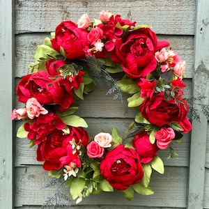 May include: A red and pink floral wreath with lush greenery. The wreath is made of silk flowers and is perfect for a spring or summer door decoration.