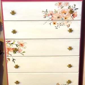 May include: A white chest of drawers with five drawers and a pink top. The drawers have gold bee-shaped knobs and floral decals. The chest of drawers is sitting on a light brown rug.