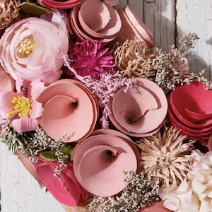 May include: Close-up of a floral wreath with a variety of pink and red flowers. The wreath includes spiral-shaped flowers, delicate blooms, and dried accents. The background is a weathered white wooden surface.