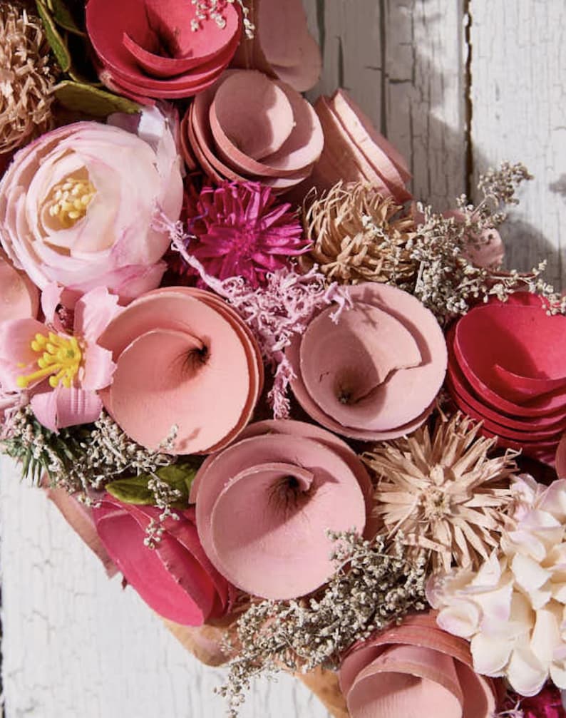 May include: Close-up of a floral wreath with a variety of pink and red flowers. The wreath includes spiral-shaped flowers, delicate blooms, and dried accents. The background is a weathered white wooden surface.