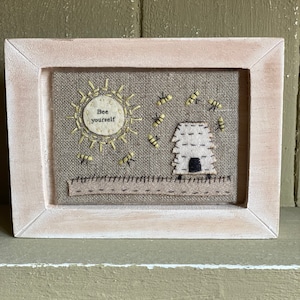 May include: A framed embroidery with a beige background featuring a yellow sun with the text "Bee yourself" and a small white beehive with black details.  There are several yellow bees flying around the beehive and sun.