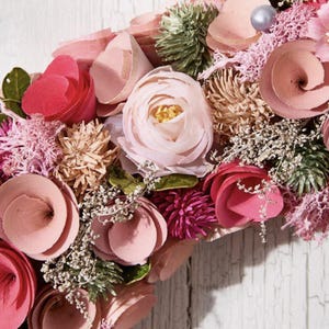 May include: A close-up of a floral wreath with a variety of pink and peach-colored flowers. The wreath includes roses, spiral-shaped blooms, and small, delicate white flowers. The background is a weathered, white wooden surface.