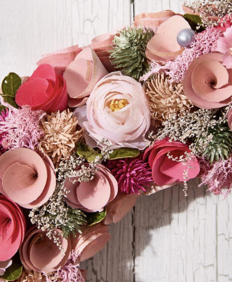 May include: A close-up of a floral wreath with a variety of pink and peach-colored flowers. The wreath includes roses, spiral-shaped blooms, and small, delicate white flowers. The background is a weathered, white wooden surface.