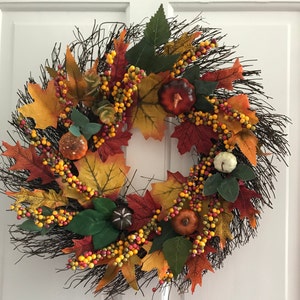 May include: A fall-themed wreath with brown twigs, orange, yellow, and red leaves, and small gourds. The wreath is decorated with small, yellow, orange, and red berries.