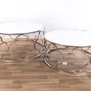 May include: Two round nesting coffee tables with a silver metal frame and white glass tops. The tables have a decorative, intricate design.